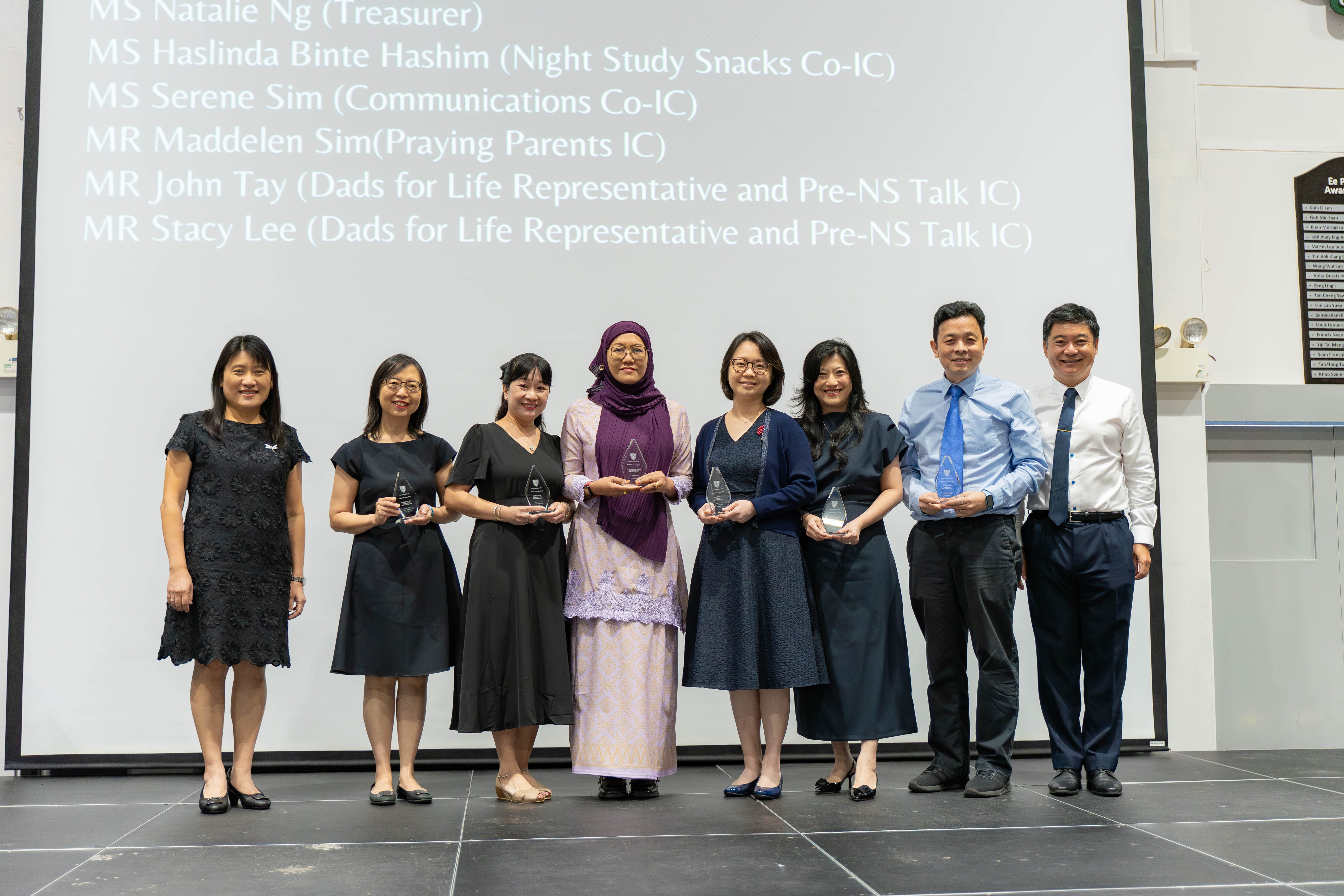 Eight people stand in front of a projected list of names, with six holding crystal award trophies.